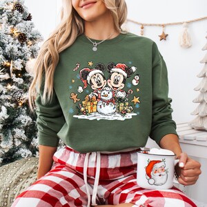 May include: Green sweatshirt featuring Mickey and Minnie Mouse in Santa hats, with a snowman, gifts, and festive accents. The design is a Christmas theme. The person is wearing red and white plaid pajama pants and holding a mug with a Santa Claus design.