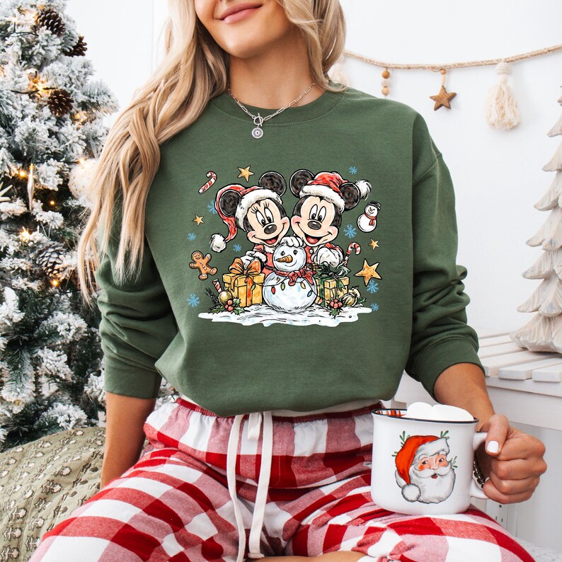 May include: Green sweatshirt featuring Mickey and Minnie Mouse in Santa hats, with a snowman, gifts, and festive accents. The design is a Christmas theme. The person is wearing red and white plaid pajama pants and holding a mug with a Santa Claus design.