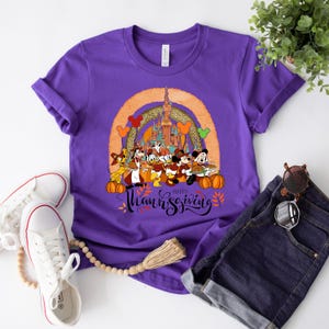 May include: A purple t-shirt featuring a graphic of Disney characters celebrating Thanksgiving. The design includes Mickey Mouse, Donald Duck, and Goofy, with a castle and the words "Happy Thanksgiving". Pumpkins and fall leaves are also included.