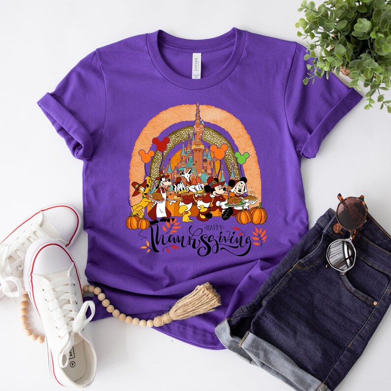 May include: A purple t-shirt featuring a graphic of Disney characters celebrating Thanksgiving. The design includes Mickey Mouse, Donald Duck, and Goofy, with a castle and the words "Happy Thanksgiving". Pumpkins and fall leaves are also included.