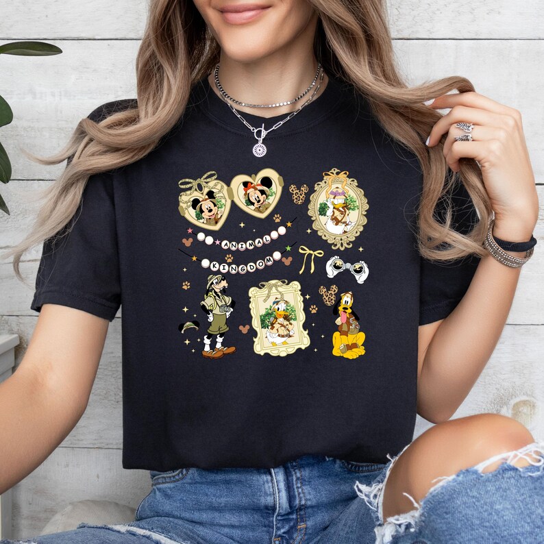 May include: Black t-shirt featuring various Disney character illustrations. Mickey and Minnie Mouse are in heart-shaped frames. Other characters include Donald Duck, Goofy, and a safari-themed Mickey Mouse. The text "Kingdom" is also visible.