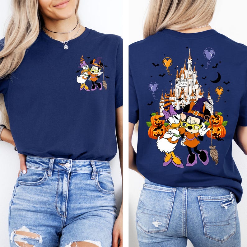 May include: Navy blue t-shirt featuring a Halloween-themed graphic of Minnie Mouse and Daisy Duck in witch costumes. The back of the shirt displays a castle, pumpkins, and balloons. The design is colorful and festive.