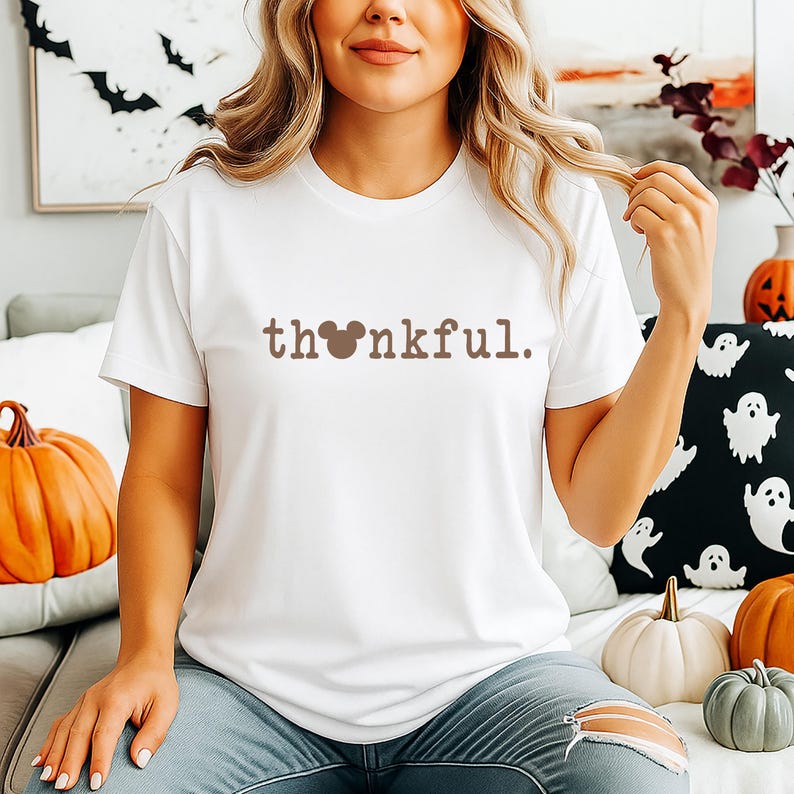 Puede incluir: Camiseta blanca con la palabra "thankful" en letras marrones. La "o" de la palabra se reemplaza con una silueta de la cabeza de Mickey Mouse. La camiseta es parte de una colecci&oacute;n con tem&aacute;tica de oto&ntilde;o o Halloween.