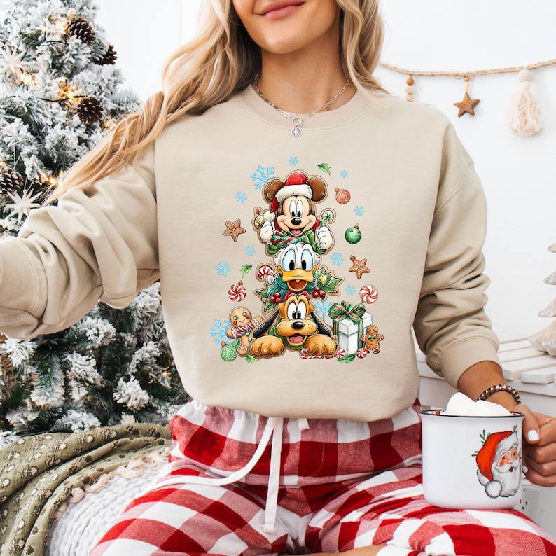 Peut inclure: Sweat-shirt beige &agrave; col rond avec un motif d'arbre de No&euml;l repr&eacute;sentant Mickey Mouse, Donald Duck et Pluto. Le motif comprend des flocons de neige, des bonhommes en pain d'&eacute;pices et des cadeaux emball&eacute;s. Le sweat-shirt est port&eacute; avec un pantalon de pyjama &agrave; carreaux rouges et blancs.