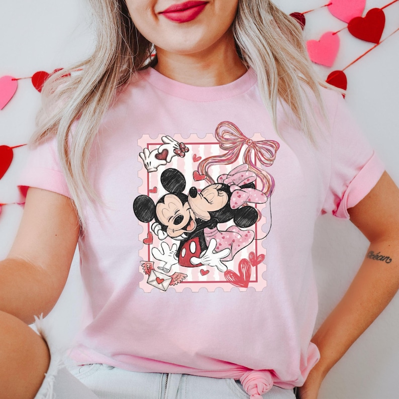 May include: Pink t-shirt featuring a graphic of Mickey and Minnie Mouse embracing within a postage stamp design. The design includes hearts, a bow, and a love letter, perfect for Valentine's Day or Disney fans.