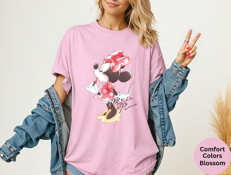 K&ouml;nnte beinhalten: Hellrosa T-Shirt mit Minnie Mouse-Grafik. Das Design zeigt Minnie Mouse in einem roten gepunkteten Kleid und Schleife. Die Worte "Minnie Mouse" sind kursiv geschrieben. Teil der Comfort Colors Blossom-Kollektion.