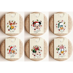May include: Six cream-colored t-shirts featuring Disney characters in festive Christmas designs. Each shirt displays a different character: Pluto, Mickey Mouse, Donald Duck, Goofy, Minnie Mouse, and Daisy Duck, all wearing holiday attire. The shirts are arranged on woven mats.