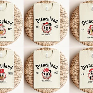 May include: Six cream-colored t-shirts with a Christmas-themed Disneyland logo featuring different Disney characters. The logo reads "Disneyland est 1955 California".