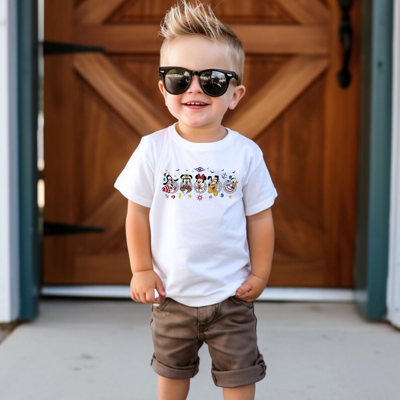 May include: A white t-shirt featuring a colorful graphic of cartoon characters. The shirt is worn by a young child wearing sunglasses and brown shorts. The graphic includes characters in various outfits.