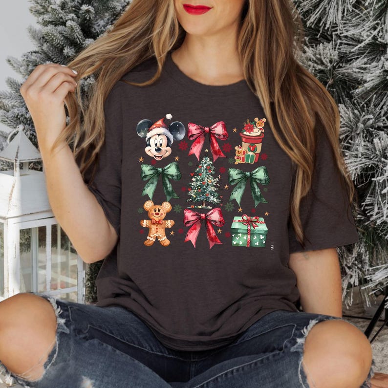 May include: A dark grey t-shirt with a Christmas-themed design featuring Mickey Mouse, a gingerbread man, a Christmas tree, bows, and gifts. The design is in a watercolor style with red, green, and gold accents.