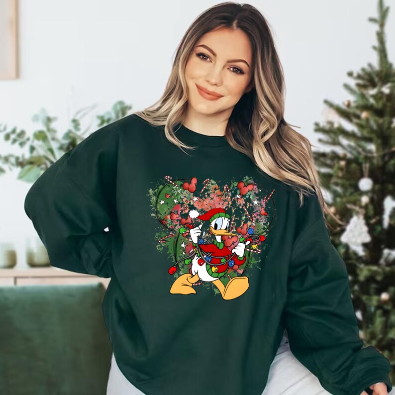 May include: A dark green sweatshirt with a Christmas-themed graphic featuring a cartoon duck wearing a Santa hat and holding Christmas lights. The duck is surrounded by green and red Christmas lights and a red and white candy cane.