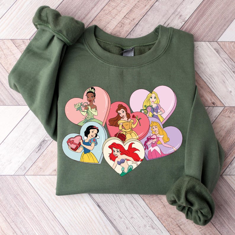 Pode incluir: Uma camisa de frio verde com uma estampa de seis princesas Disney em molduras em forma de cora&ccedil;&atilde;o. As princesas s&atilde;o Branca de Neve, Cinderela, Ariel, Bela, Aurora e Tiana.