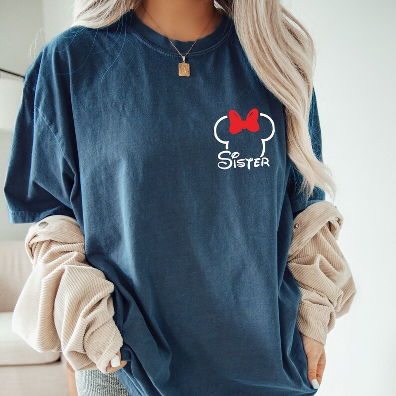 Puede incluir: Una camiseta azul marino con un contorno blanco de una cabeza de rat&oacute;n de dibujos animados con un lazo rojo y la palabra "Hermana" debajo.