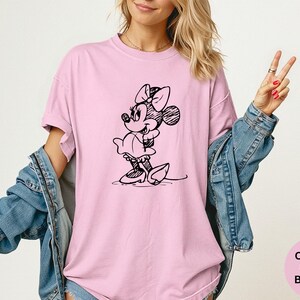 May include: A light pink t-shirt featuring a black sketch of Minnie Mouse. The shirt has short sleeves and a crew neck. The text "Comfort Colors Blossom" is in a pink circle in the lower right corner.