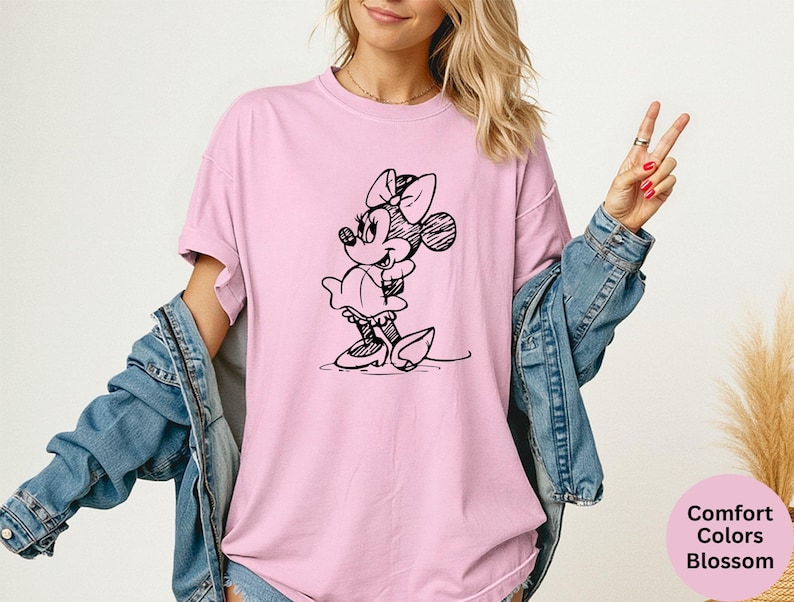 May include: A light pink t-shirt featuring a black sketch of Minnie Mouse. The shirt has short sleeves and a crew neck. The text "Comfort Colors Blossom" is in a pink circle in the lower right corner.