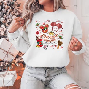 May include: White sweatshirt with a festive design featuring a Mickey Mouse gingerbread cookie, a Minnie Mouse bow headband, and a hot chocolate. The phrase "'Tis the Season" is written in a circular pattern with holiday accents.