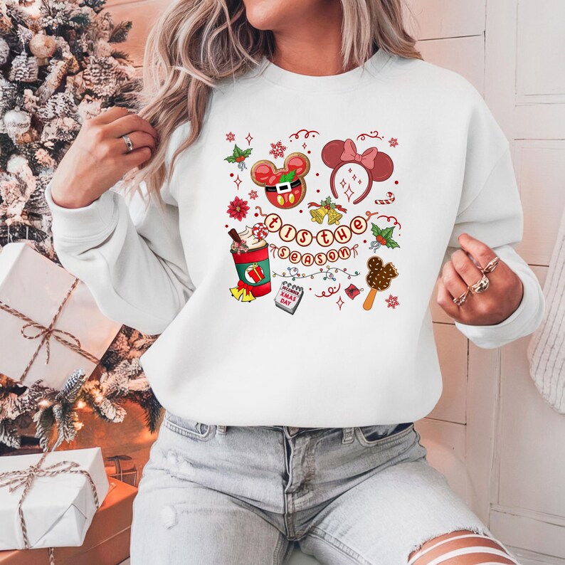 May include: White sweatshirt with a festive design featuring a Mickey Mouse gingerbread cookie, a Minnie Mouse bow headband, and a hot chocolate. The phrase "'Tis the Season" is written in a circular pattern with holiday accents.