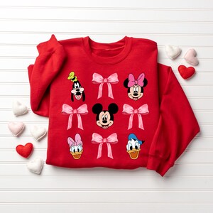 May include: Red sweatshirt with a festive Valentine's Day theme featuring Mickey Mouse, Minnie Mouse, Donald Duck, Daisy Duck, and Goofy. Each character is wearing a pink bow.