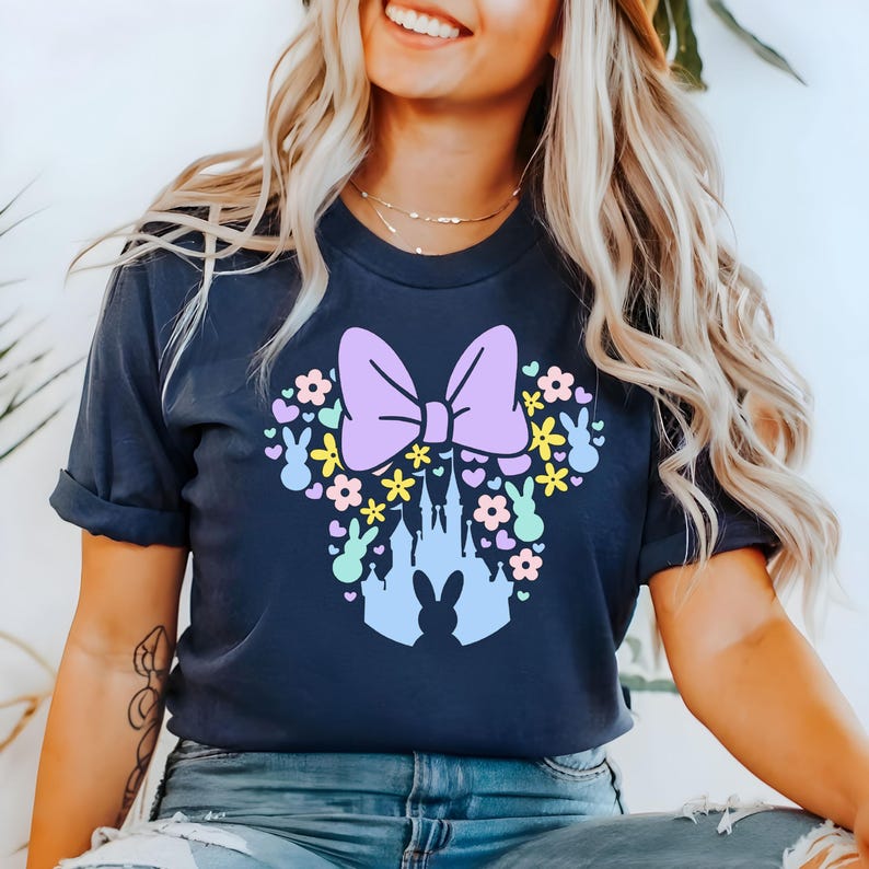 May include: Navy blue t-shirt featuring a design of a purple bow, flowers, bunnies, and a castle silhouette. The design is arranged in the shape of a mouse head. The shirt is a comfortable, casual style.