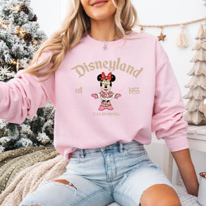 May include: A light pink Disneyland sweatshirt featuring a Minnie Mouse graphic. The graphic includes the text "Disneyland" above a Minnie Mouse illustration, with "est 1955" and "CALIFORNIA" below. The sweatshirt has long sleeves.