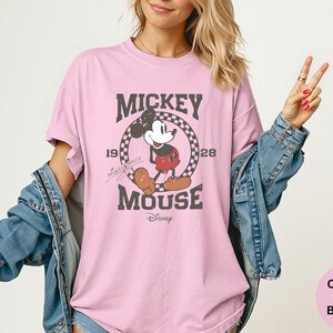 May include: A light pink t-shirt featuring a distressed graphic of Mickey Mouse with the text "Mickey Mouse" and the years "1928" and "1928". The shirt is part of the Comfort Colors Blossom collection.