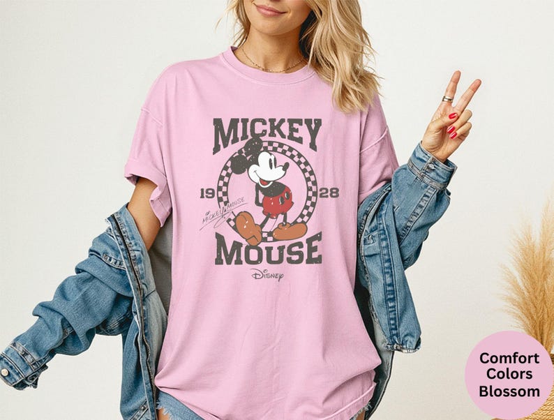 May include: A light pink t-shirt featuring a distressed graphic of Mickey Mouse with the text "Mickey Mouse" and the years "1928" and "1928". The shirt is part of the Comfort Colors Blossom collection.