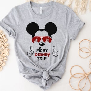 Op de afbeelding: Grijs T-shirt met een zwart-witte Mickey Mouse die een rode zonnebril draagt met een kasteel silhouet. De tekst "First Disney Trip" staat onder Mickey Mouse.