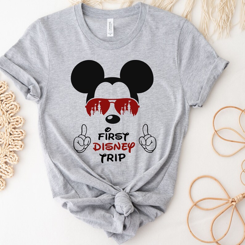 Op de afbeelding: Grijs T-shirt met een zwart-witte Mickey Mouse die een rode zonnebril draagt met een kasteel silhouet. De tekst "First Disney Trip" staat onder Mickey Mouse.