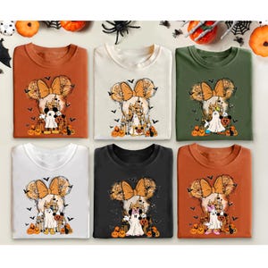May include: Six different t-shirts with a Halloween-themed design featuring a cartoon character with a bow, a castle, and pumpkins. The shirts are in various colors, including orange, white, black, and green.