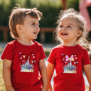 Op de afbeelding: Twee kinderen dragen rode T-shirts met een kleurrijke afbeelding van Assepoester's kasteel en Mickey Mouse-oren. De afbeelding bevat vuurwerk en andere details.