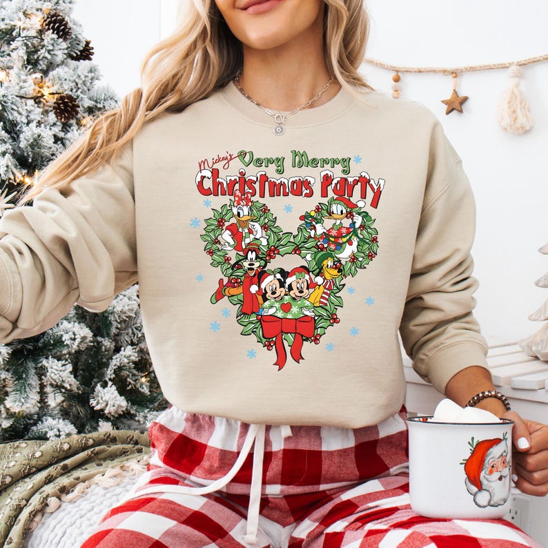 Op de afbeelding: Beige sweatshirt met een kerstthema. De afbeelding toont Mickey Mouse en vrienden, met de tekst "Mickey's Very Merry Christmas Party". Het ontwerp is hartvormig, met sneeuwvlokken en een rode strik.