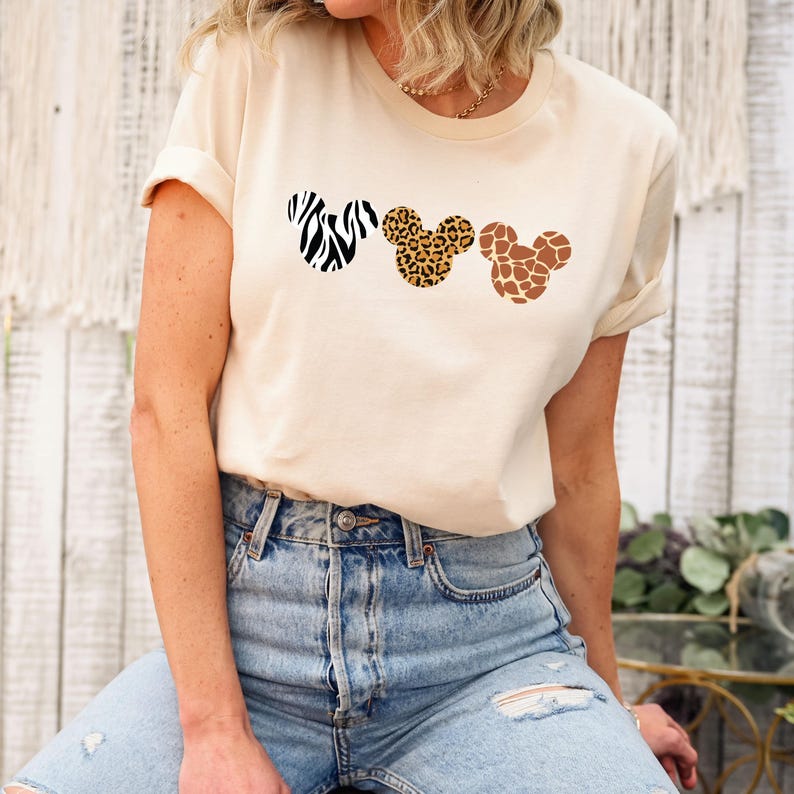 May include: A cream-colored t-shirt featuring three Mickey Mouse head silhouettes with animal print patterns. The patterns include zebra stripes, leopard spots, and giraffe spots. The shirt has rolled-up sleeves and is worn with blue jeans.
