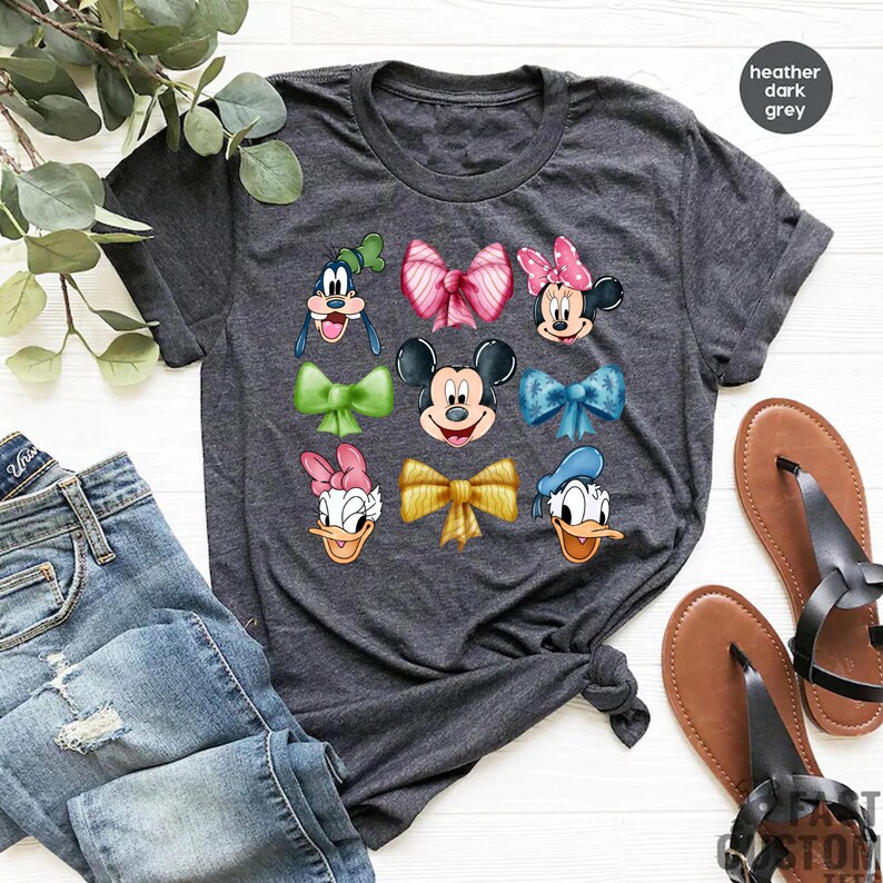May include: A heather dark grey t-shirt with a graphic print of Disney characters wearing bows. The characters include Mickey Mouse, Minnie Mouse, Donald Duck, Daisy Duck, Goofy, and Pluto.