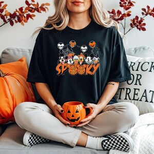 May include: Black t-shirt with a Halloween graphic featuring cartoon characters, balloons, and the word "Spooky" in orange. An orange pumpkin mug is held in the lap. The background includes autumn foliage and a decorative pillow.