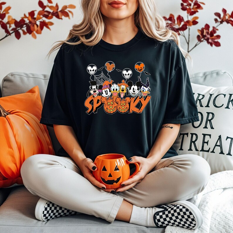 May include: Black t-shirt with a Halloween graphic featuring cartoon characters, balloons, and the word "Spooky" in orange. An orange pumpkin mug is held in the lap. The background includes autumn foliage and a decorative pillow.