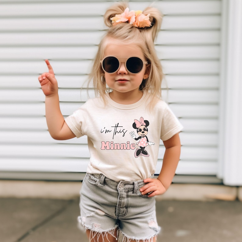 Puede incluir: Una ni&ntilde;a peque&ntilde;a con una camiseta blanca con un gr&aacute;fico de Minnie Mouse rosa y el texto "i'm this Minnie". Tambi&eacute;n lleva pantalones cortos de mezclilla y gafas de sol.