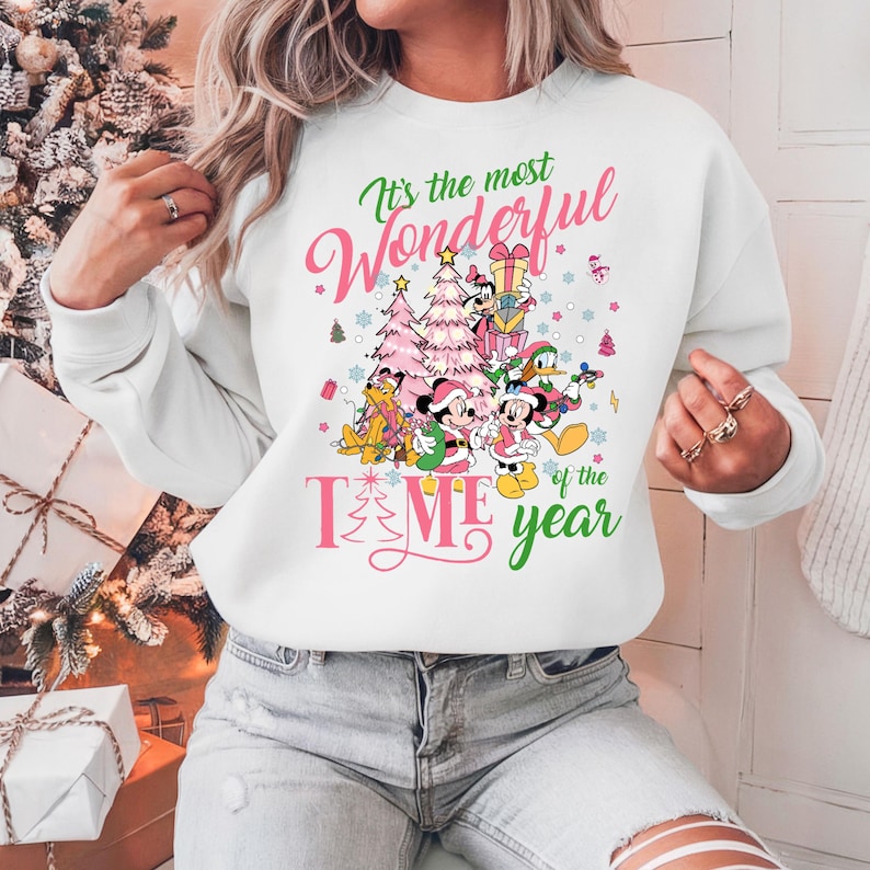 May include: White sweatshirt with a pink and green Christmas design featuring Mickey Mouse and friends. The text on the sweatshirt reads "It's the most Wonderful Time of the Year."