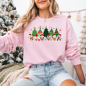 May include: A light pink sweatshirt featuring a festive Christmas design. The graphic includes cartoon characters wearing Santa hats and Christmas trees. The design is colorful and cheerful, perfect for the holiday season.