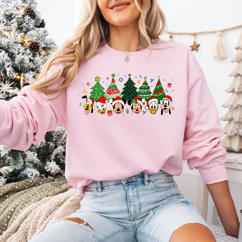 May include: A light pink sweatshirt featuring a festive Christmas design. The graphic includes cartoon characters wearing Santa hats and Christmas trees. The design is colorful and cheerful, perfect for the holiday season.