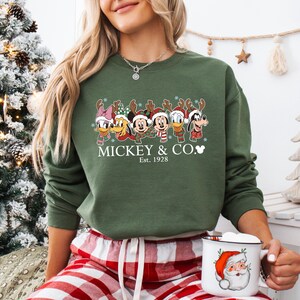 K&ouml;nnte beinhalten: Olivgr&uuml;nes Sweatshirt mit einem weihnachtlichen Disney-Design. Das Design zeigt Mickey Mouse, Donald Duck und andere Charaktere mit Rentiergeweihen. Der Text "Mickey & Co. Est. 1928" ist darunter aufgedruckt. Eine Tasse mit Santa Claus-Design wird gehalten.
