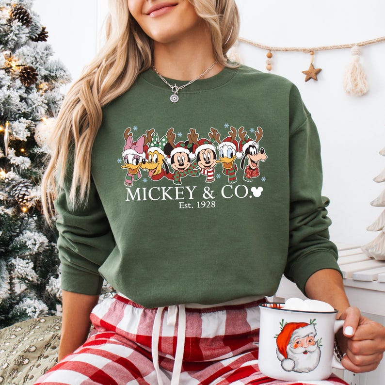 K&ouml;nnte beinhalten: Olivgr&uuml;nes Sweatshirt mit einem weihnachtlichen Disney-Design. Das Design zeigt Mickey Mouse, Donald Duck und andere Charaktere mit Rentiergeweihen. Der Text "Mickey & Co. Est. 1928" ist darunter aufgedruckt. Eine Tasse mit Santa Claus-Design wird gehalten.