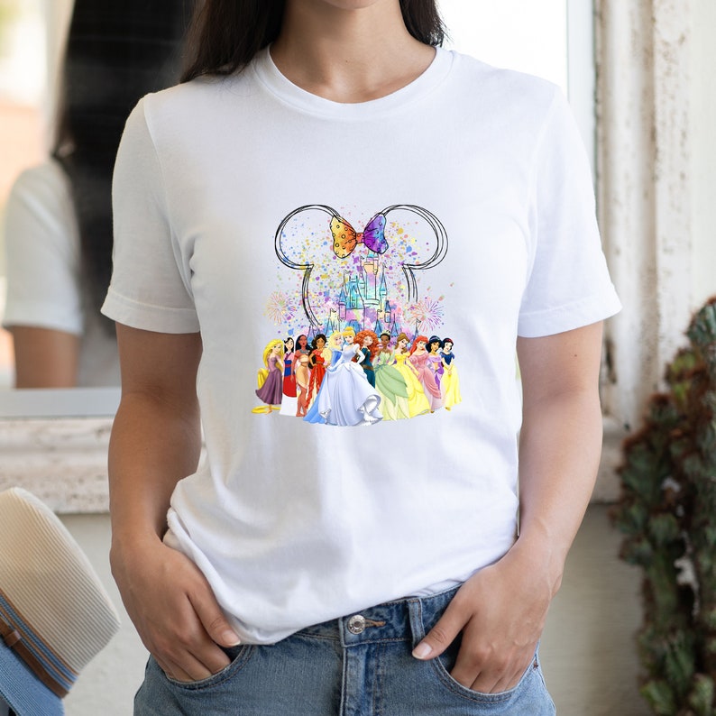 Puede incluir: Camiseta blanca con un gr&aacute;fico colorido de princesas Disney de pie frente al castillo de Cenicienta. El gr&aacute;fico incluye orejas de Mickey Mouse con un lazo y fuegos artificiales.