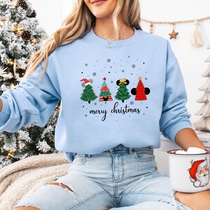 May include: Light blue sweatshirt with a festive Christmas tree design. The graphic features four stylized Christmas trees with mouse ears and a Santa hat, with the words "merry christmas" below. The sweatshirt is a solid light blue color.