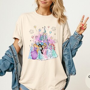 K&ouml;nnte beinhalten: Elfenbeinfarbenes T-Shirt mit einer farbenfrohen Illustration mehrerer Prinzessinnen vor einem Schloss. Die Grafik enth&auml;lt Feuerwerk und Luftballons. Das Shirt ist von der Marke Comfort Colors.