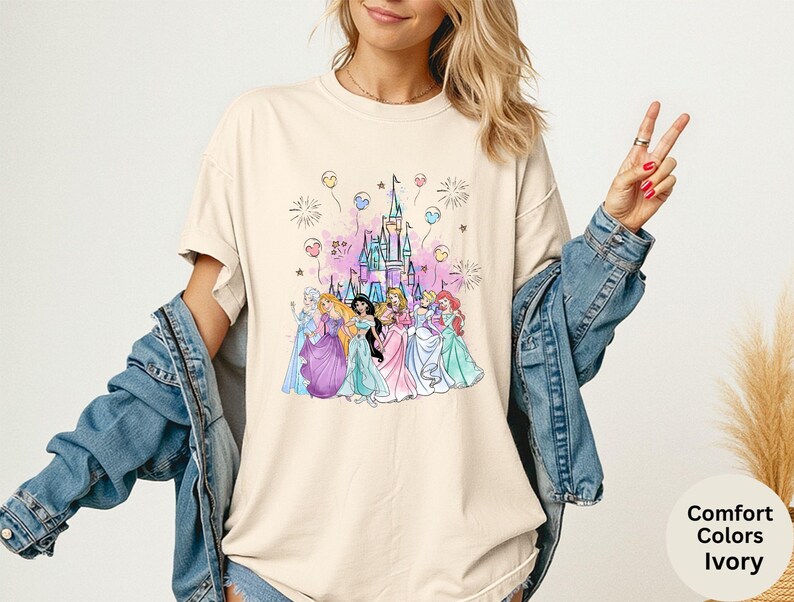 K&ouml;nnte beinhalten: Elfenbeinfarbenes T-Shirt mit einer farbenfrohen Illustration mehrerer Prinzessinnen vor einem Schloss. Die Grafik enth&auml;lt Feuerwerk und Luftballons. Das Shirt ist von der Marke Comfort Colors.