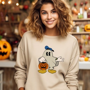 Puede incluir: Una sudadera beige con un fantasma de dibujos animados que lleva un gorro de marinero azul y sostiene una calabaza tallada. El fantasma tiene una expresi&oacute;n de sorpresa y lleva una s&aacute;bana blanca.