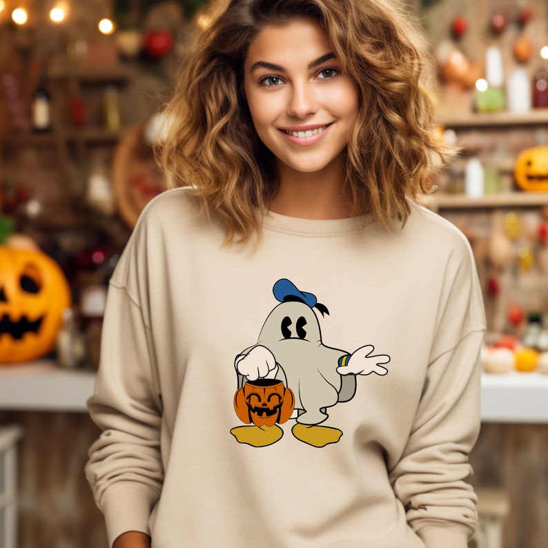 Puede incluir: Una sudadera beige con un fantasma de dibujos animados que lleva un gorro de marinero azul y sostiene una calabaza tallada. El fantasma tiene una expresi&oacute;n de sorpresa y lleva una s&aacute;bana blanca.