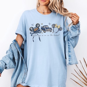 May include: Pale blue t-shirt with a graphic of a blue car, mouse ears, and the text "Racing is in the heart." The design includes the words "Fabulous Hudson Hornet". The shirt has a loose fit.