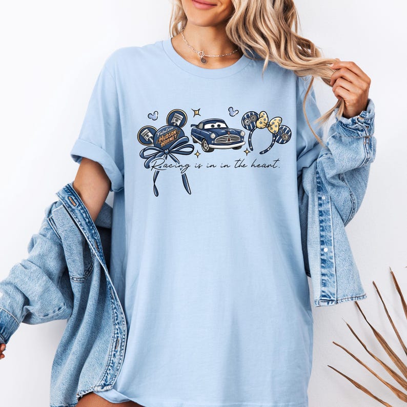 May include: Pale blue t-shirt with a graphic of a blue car, mouse ears, and the text "Racing is in the heart." The design includes the words "Fabulous Hudson Hornet". The shirt has a loose fit.