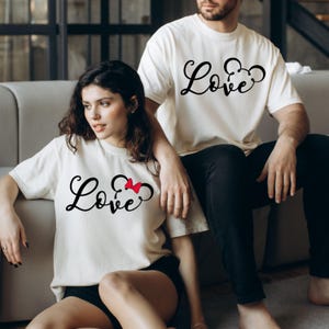 K&ouml;nnte beinhalten: Ein Paar tr&auml;gt wei&szlig;e T-Shirts mit dem Wort "Love" und einer Mickey-Maus-Silhouette. Das Shirt der Frau hat eine rote Schleife am Mickey-Maus-Ohr.